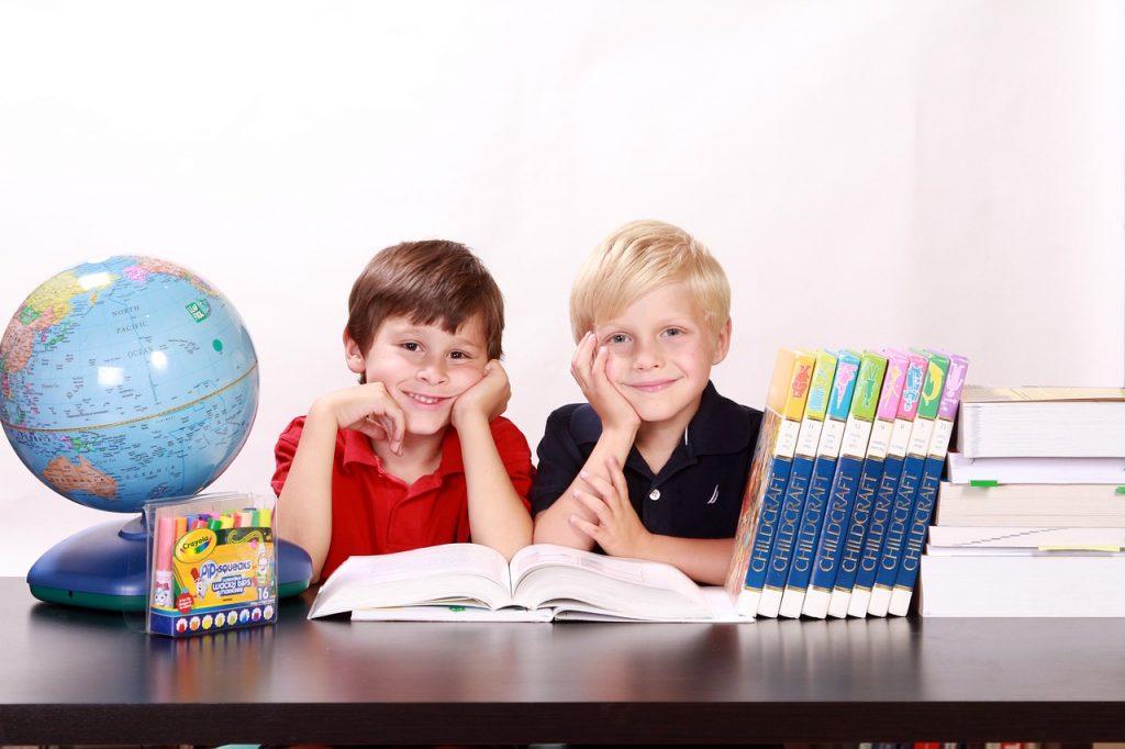 Two boys smiling at the camera resting their faces in their hands. A globe is on their left and books to their right. An open text book is in front of them.