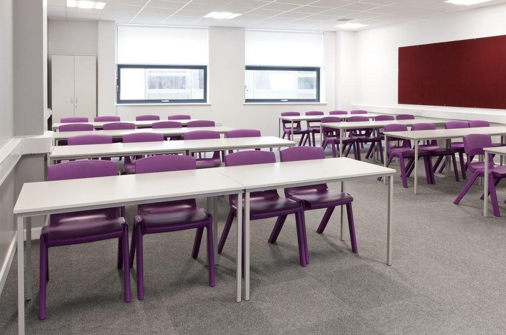 A photograph of an empty classroom. The chairs are purple, the walls and desks are white.